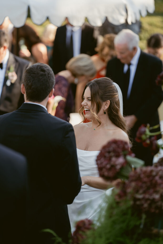Bride smiles brightly at the groom during their outdoor wedding ceremony surrounded by loved ones.