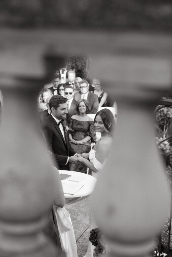 Black and white photo of bride and groom exchanging vows, seen through stone railing during ceremony.