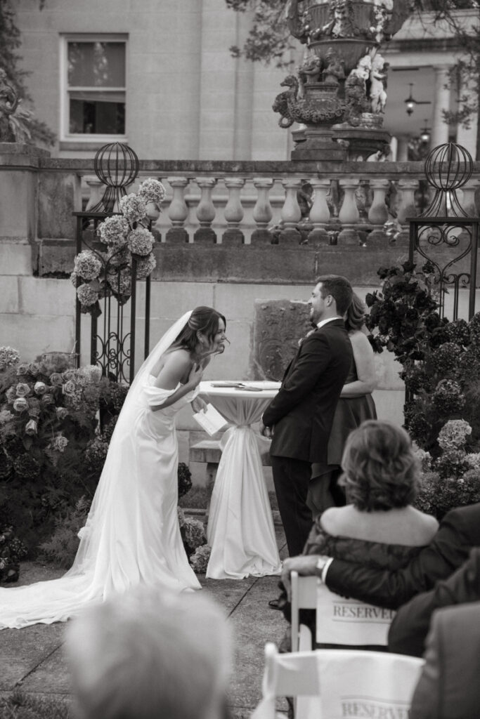 Black and white photo of bride laughing during wedding ceremony while holding groom’s hands at the altar.