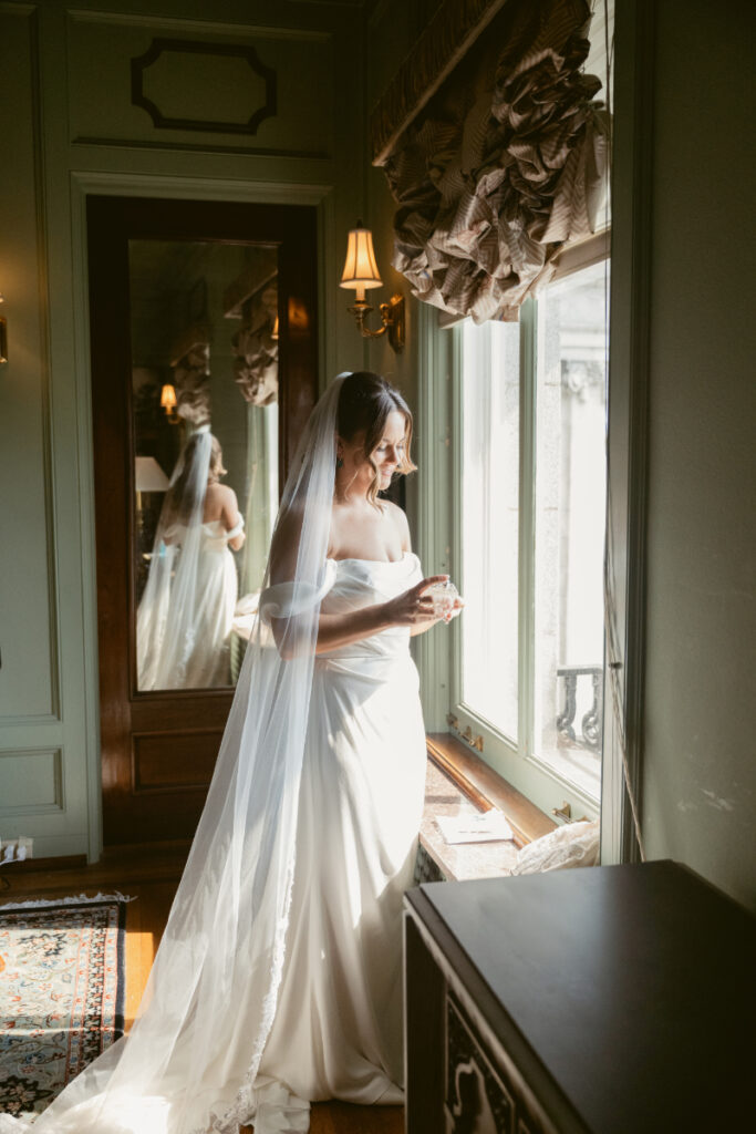 Bride smiling by the window while applying perfume before her Laurel Court wedding ceremony.