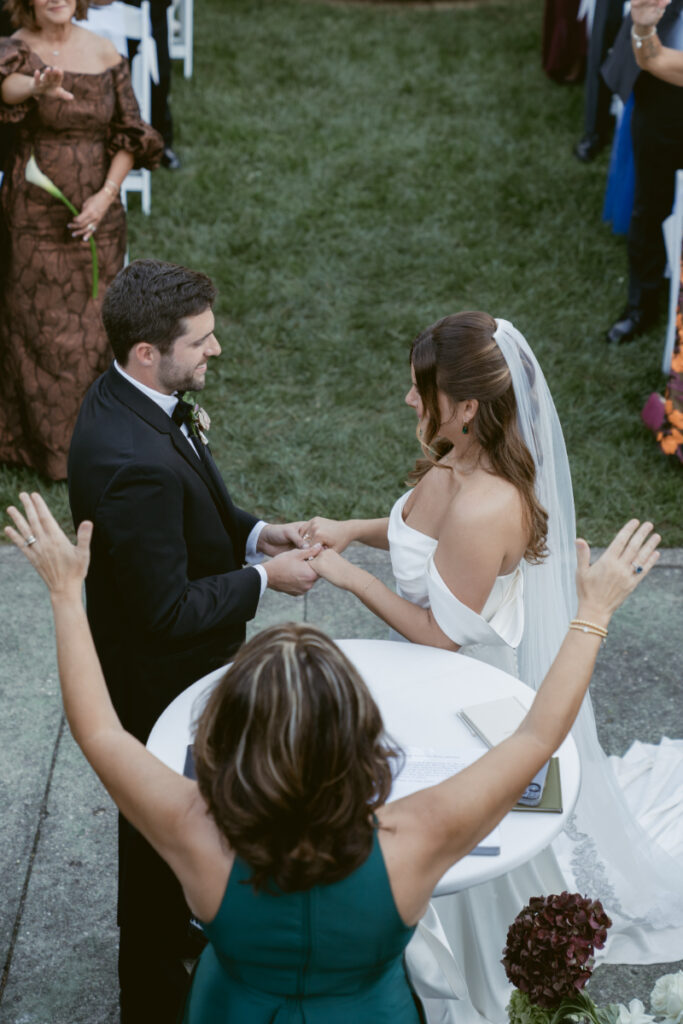 Bride and groom hold hands during outdoor wedding ceremony as officiant raises arms to celebrate.