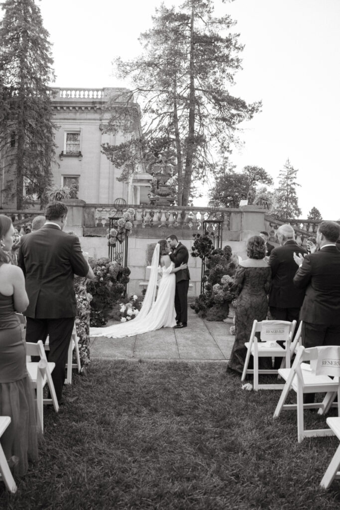 Bride and groom share a kiss at the altar as wedding guests applaud during outdoor ceremony.