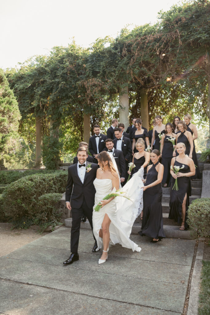 The bride and groom lead their wedding party down the garden steps at Laurel Court in Cincinnati, surrounded by bridesmaids in elegant black dresses and groomsmen in classic tuxedos.
