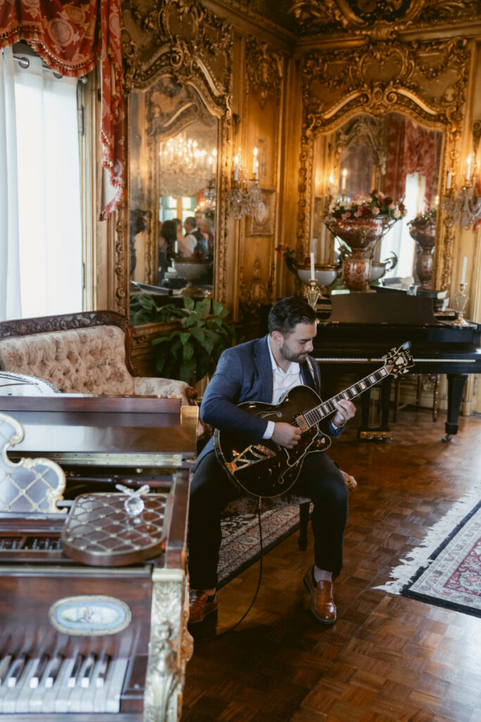 Man playing electric guitar in ornate, European-style room with gilded walls, mirrors, and antique furniture.