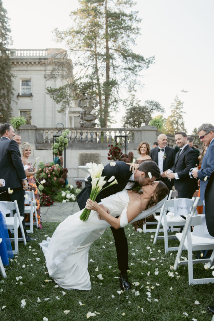 Groom dips and kisses bride during joyful wedding ceremony recessional surrounded by guests and flower petals.