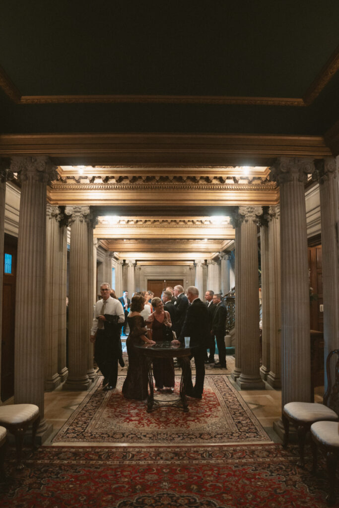 Guests gather and mingle in an ornate hallway with grand columns and rich decorative details.