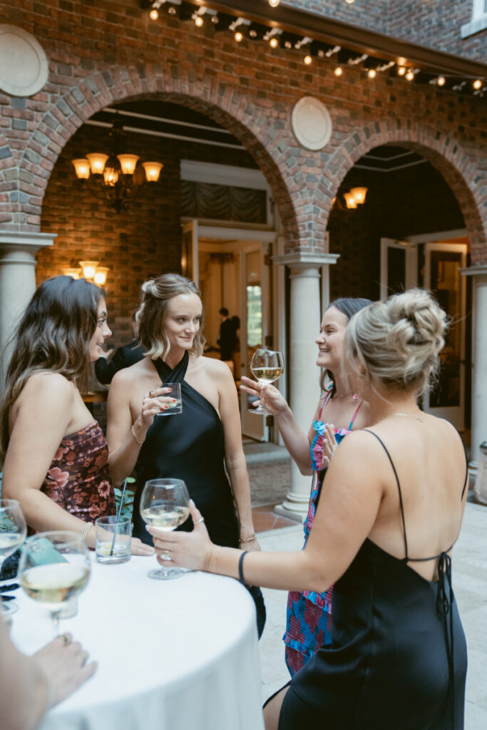 Four women in cocktail attire laugh and chat while holding drinks during an outdoor wedding reception.