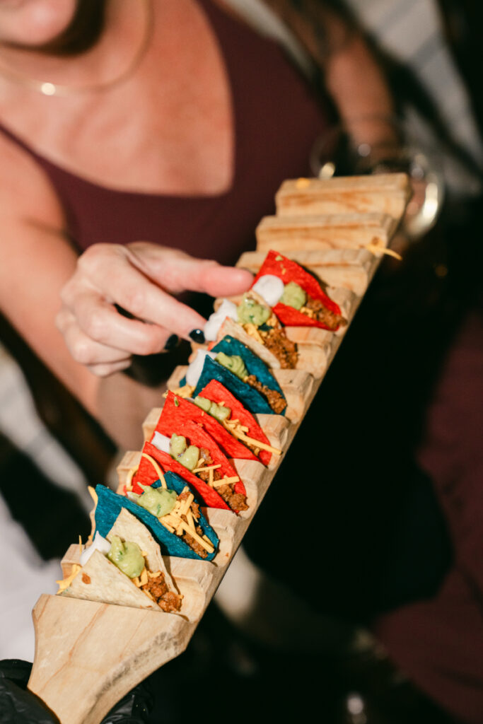 Guest picks up a colorful mini taco from a wooden tray of vibrant hors d'oeuvres at reception.