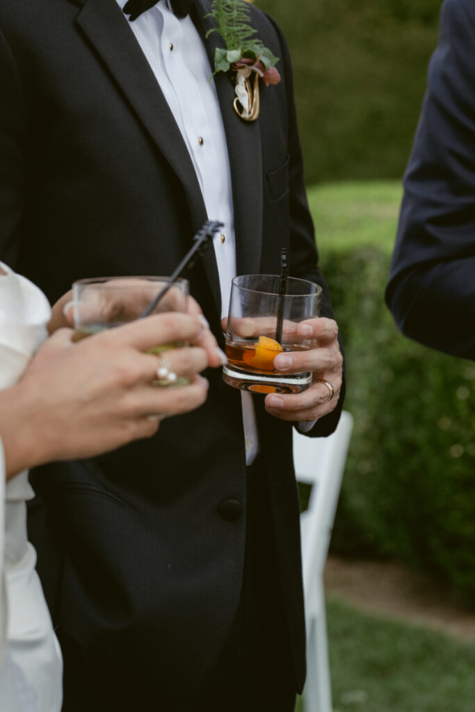 Close-up of couple holding cocktails in lowball glasses during an outdoor wedding celebration in formal attire.