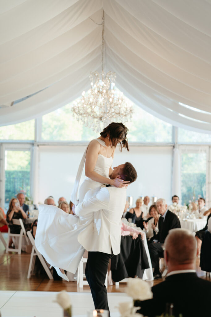 The groom lifts the bride mid-spin during their first dance, surrounded by smiling guests and candlelight.