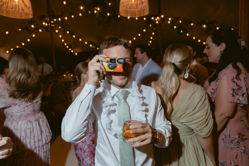 A guest in party mode snaps a photo with a disposable camera, drink in hand and smile ready.