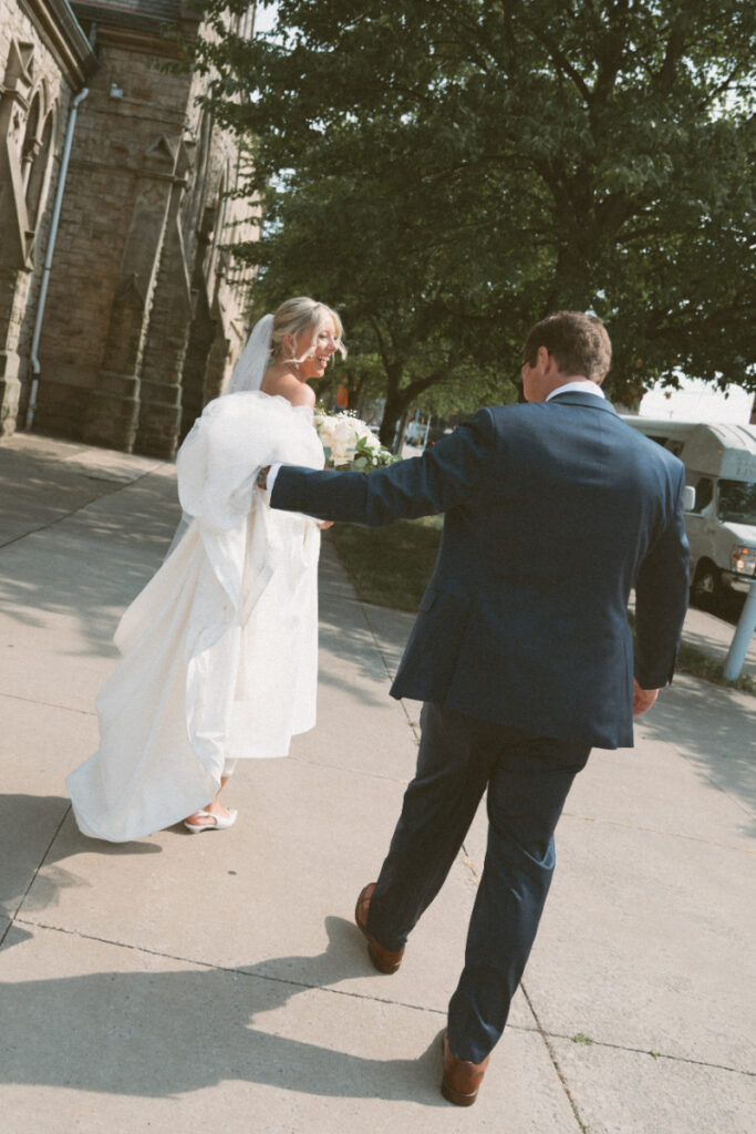 The bride laughs joyfully as she walks arm-in-arm with her father outside the church on a sunny day.