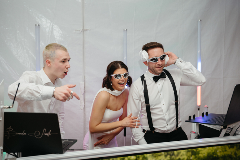 The bride and groom join the DJ booth in sunglasses, laughing and hyping the party with full energy.