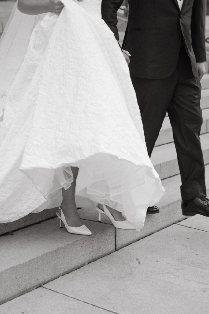 A close-up of the bride’s heels and gown as she steps carefully down stone stairs beside her escort.