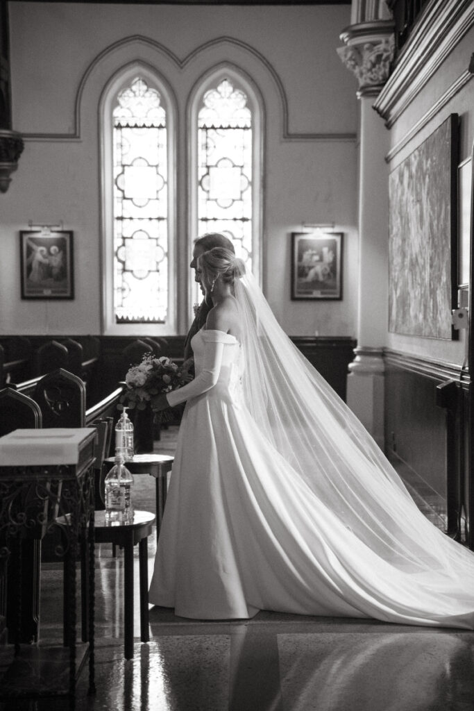The bride stands in stillness inside a sunlit chapel, her long veil trailing behind her gracefully.