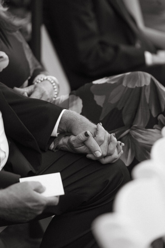 An older couple sits hand in hand during the ceremony, quietly sharing the weight of the moment.