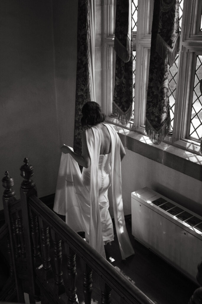 The bride walks down a quiet staircase, bathed in soft window light, dress gathered gently in hand.