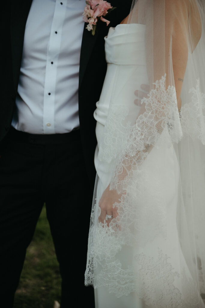 Close-up of bride and groom in wedding attire, featuring lace veil and classic black tuxedo.