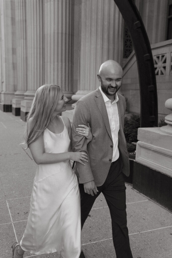 Couple walking arm in arm and smiling in front of a historic building with tall columns.