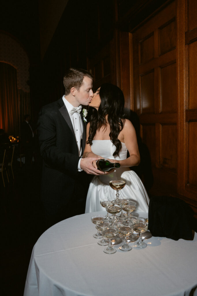 The couple shares a kiss while pouring champagne into a tower at their cozy Christmas wedding.