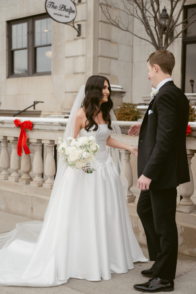 Bride and groom smile outside The Phelps, dressed for a winter wedding with red bow decor.