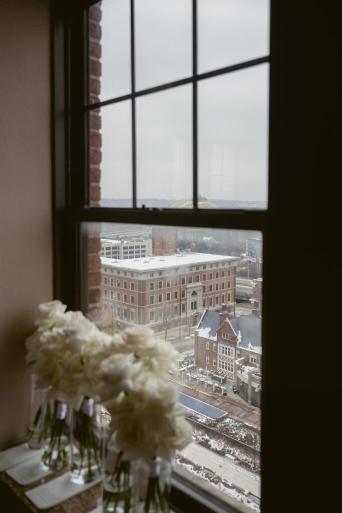 Snowy city view from hotel window on cozy Christmas wedding day, with white roses in foreground.