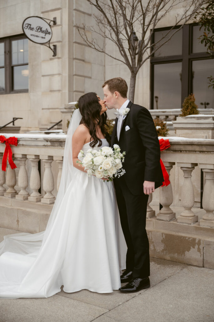 Bride and groom share a kiss outside The Phelps, surrounded by festive red holiday bows.