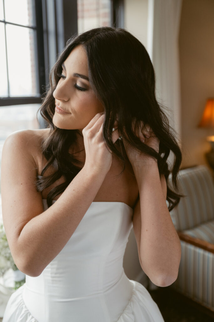 Bride adjusting earrings in natural light before cozy Christmas wedding, wearing strapless satin gown.