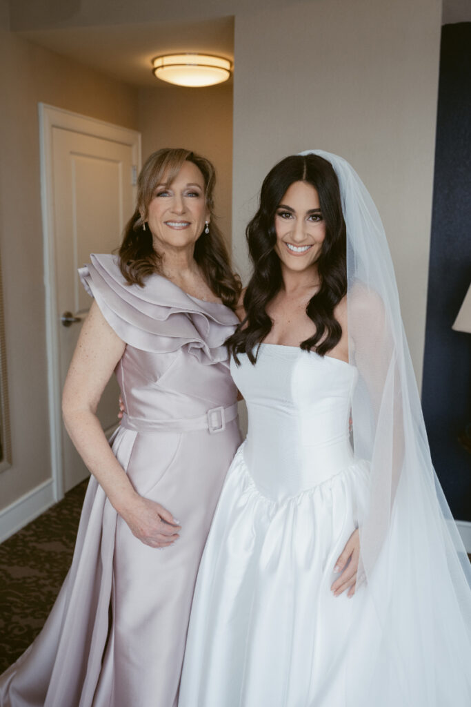 Bride and mother smiling together before cozy Christmas wedding, both dressed in elegant gowns.