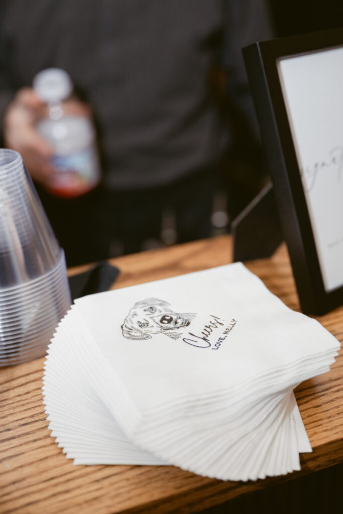 A stack of personalized cocktail napkins featuring a cute illustration of a dog named Chevy and the phrase “Chevy approves,” sitting at a bar setup with clear plastic cups.