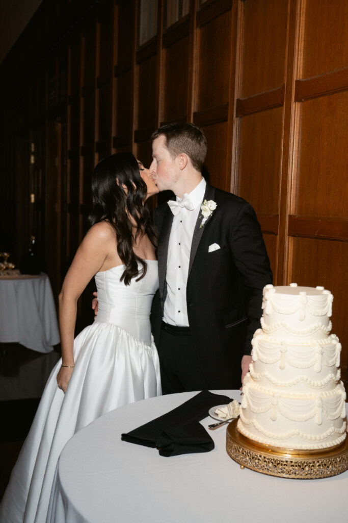 The couple shares a kiss beside their elegant tiered cake at their winter wedding reception.