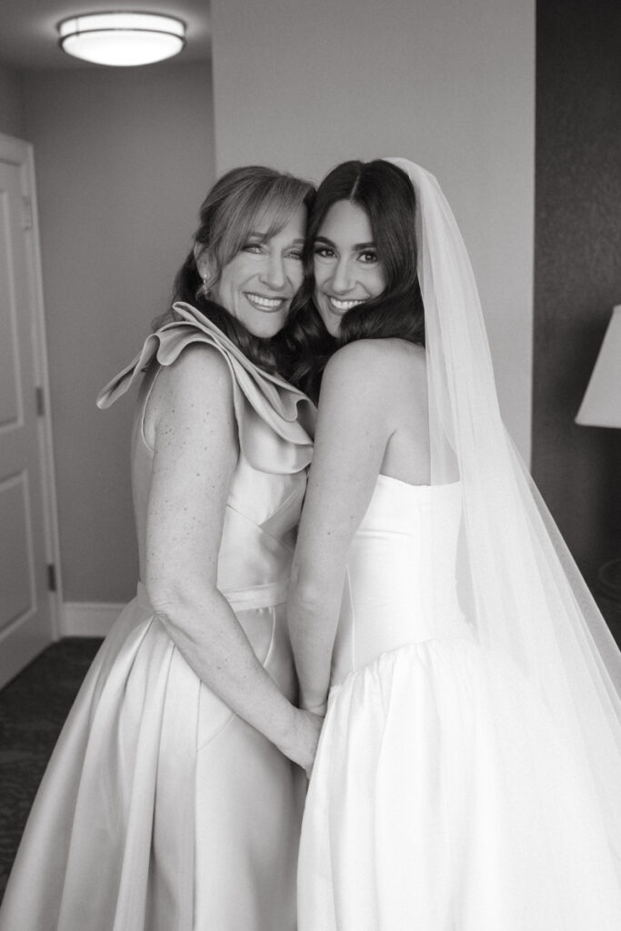 Black and white photo of bride and mother hugging before cozy Christmas wedding, smiling at camera.