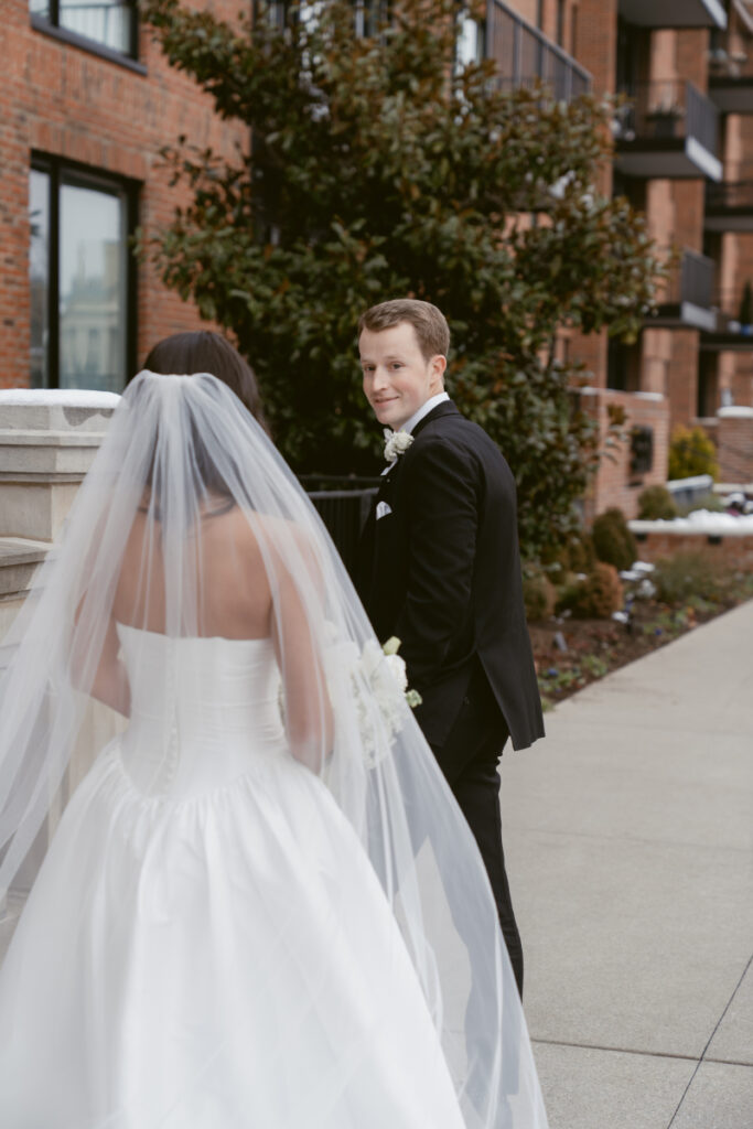 Groom glances back at the bride with a smile as they walk together on a city sidewalk.