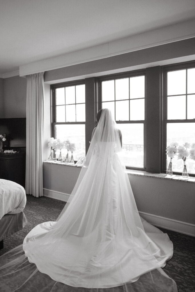 Black and white photo of bride in gown and veil facing window before cozy Christmas wedding day.