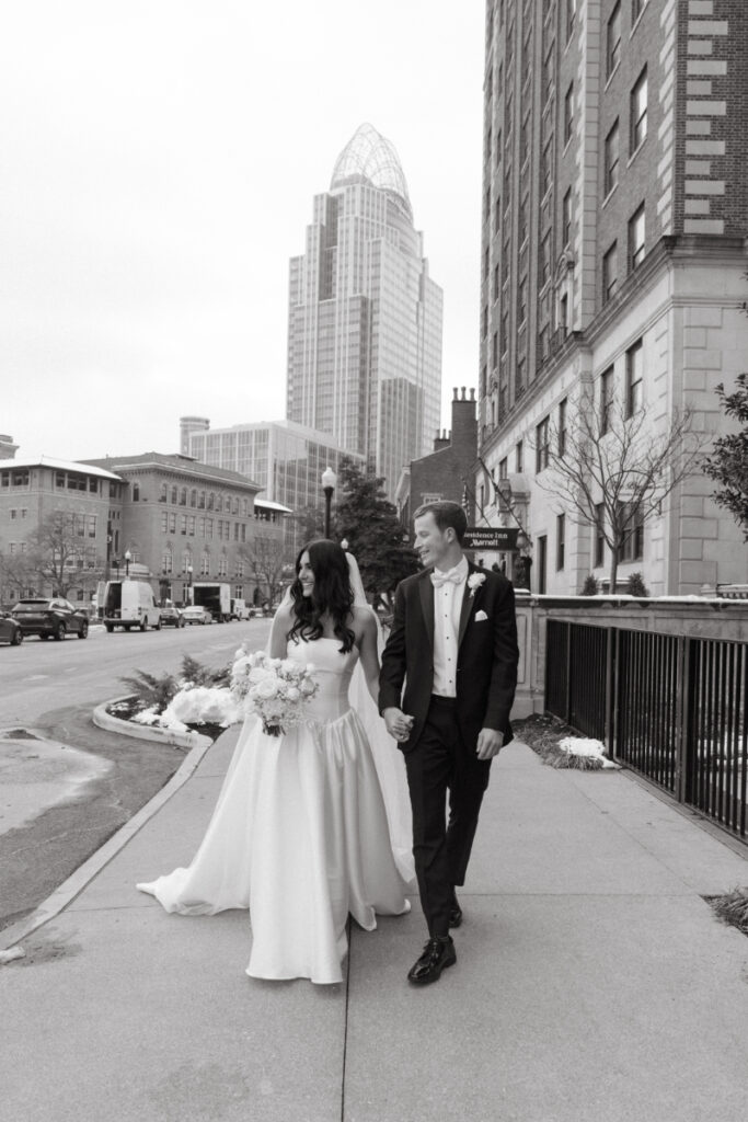 Bride and groom walk hand in hand downtown with city skyline in the background.