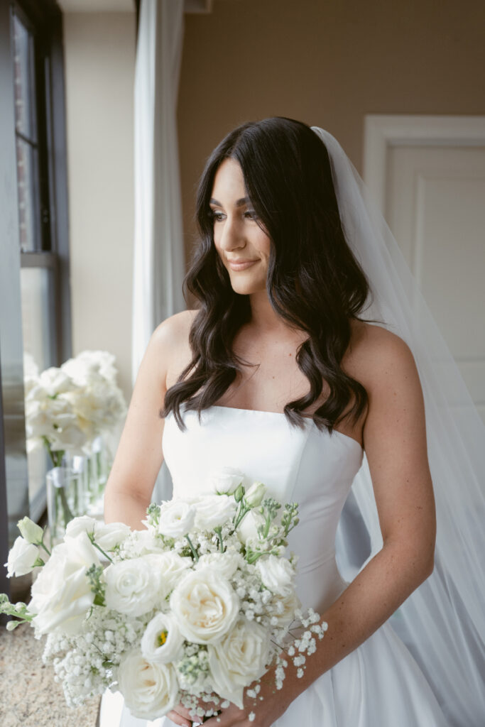 Bride holding white rose bouquet and looking out window before cozy Christmas wedding ceremony.