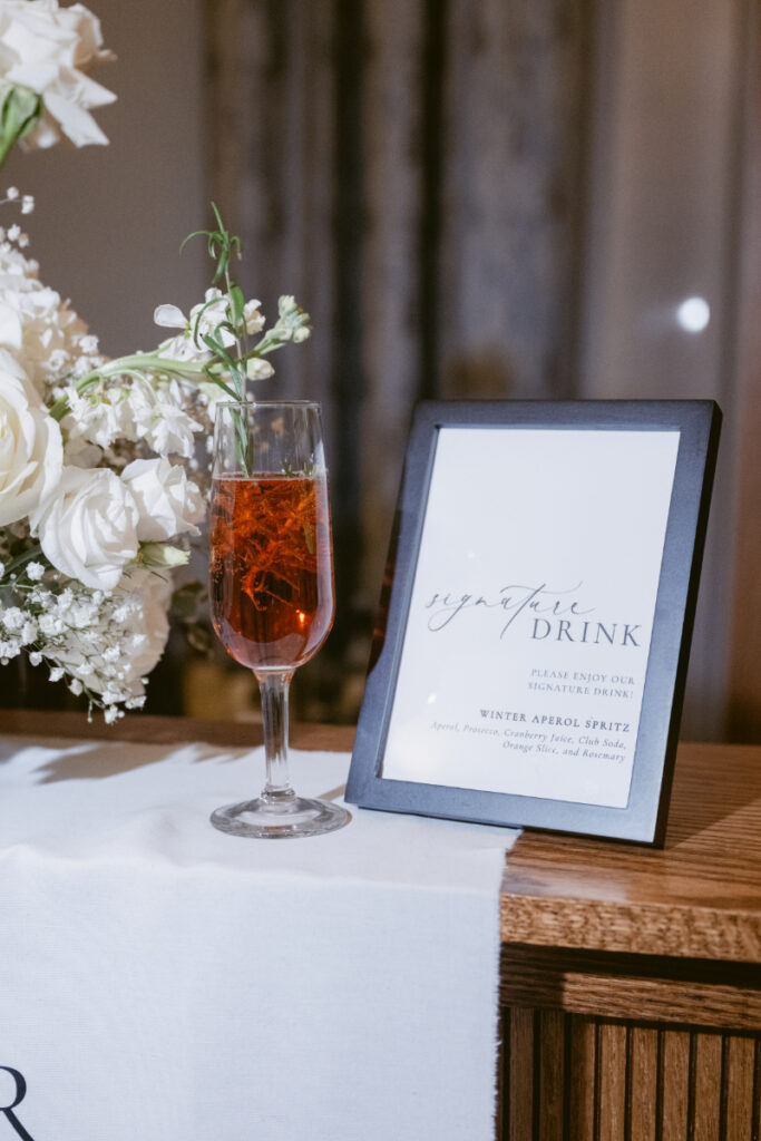 A beautifully styled bar area showing a signature drink, framed sign and a glass of the pink drink surrounded by white flowers.