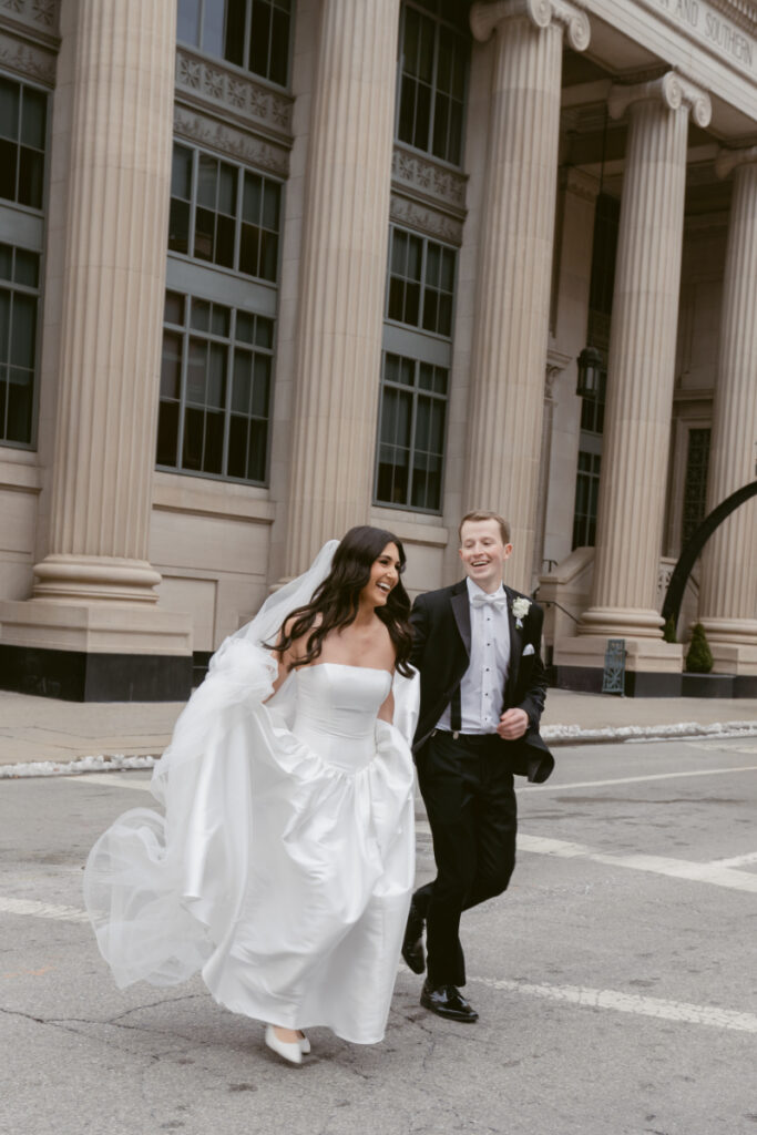 Bride and groom joyfully run across the street in front of grand columns, laughing together.