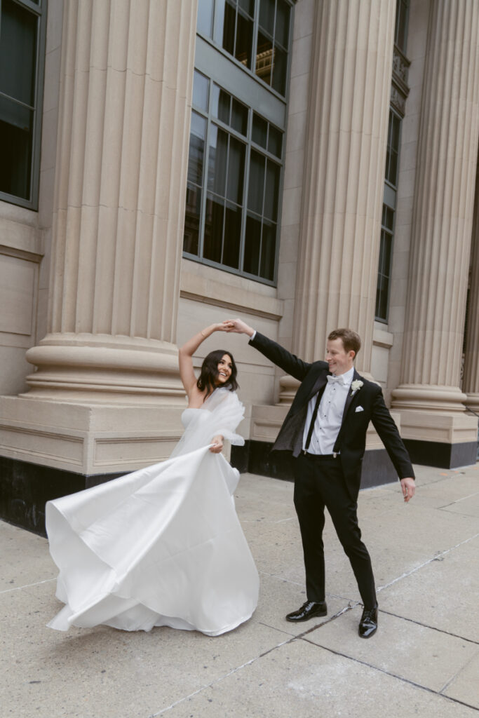 Groom twirls bride on the sidewalk as her dress flows in front of tall city columns.