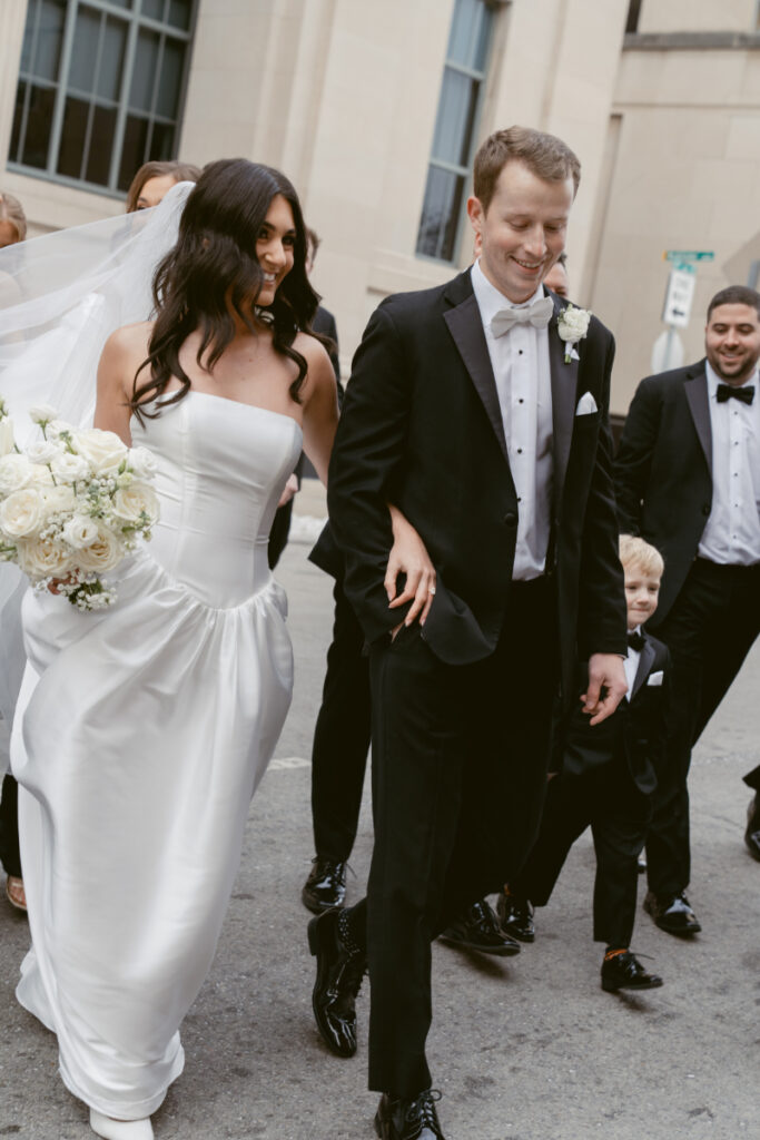 Bride and groom walk hand in hand, smiling, surrounded by wedding party on city street.