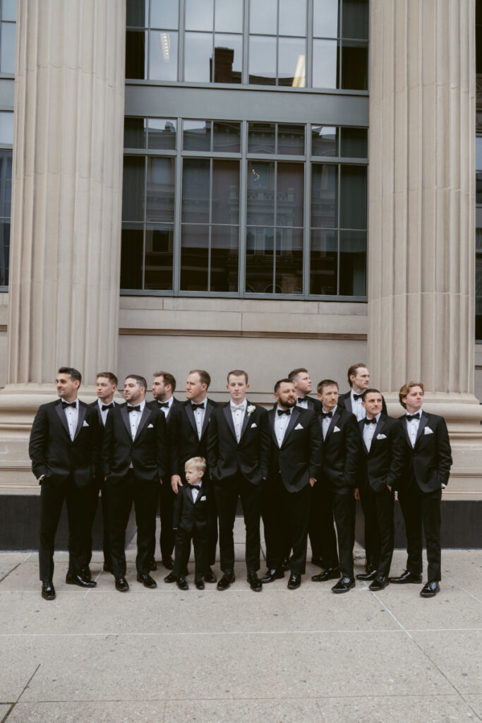 The groom stands with his groomsmen and ring bearer in front of a grand building.