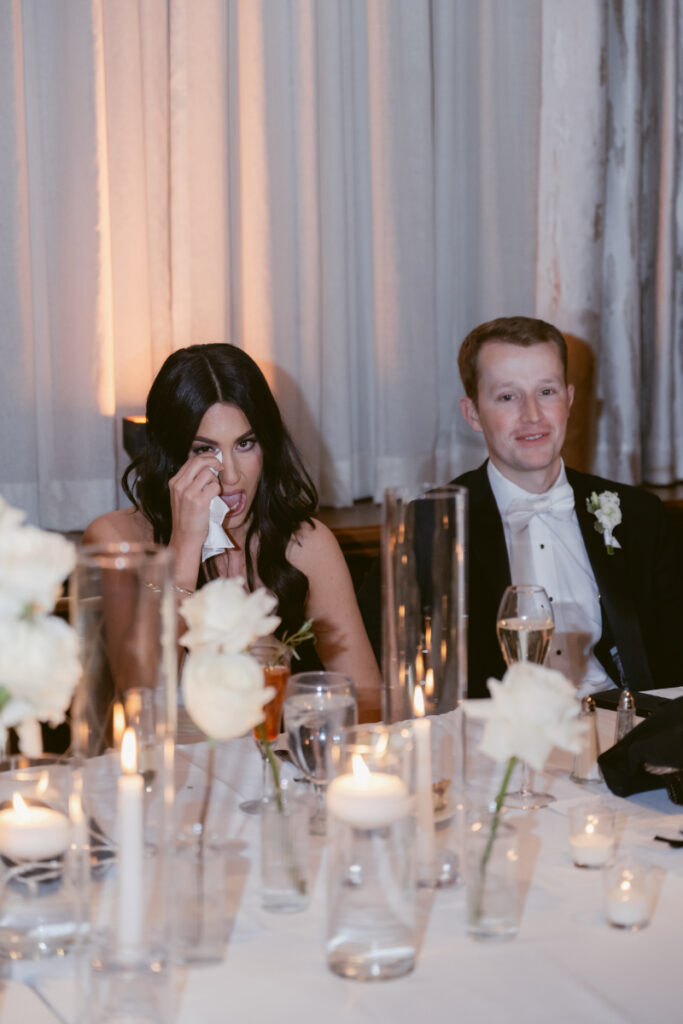 The bride wipes away tears during a heartfelt toast, seated beside the groom at their cozy Christmas wedding reception.