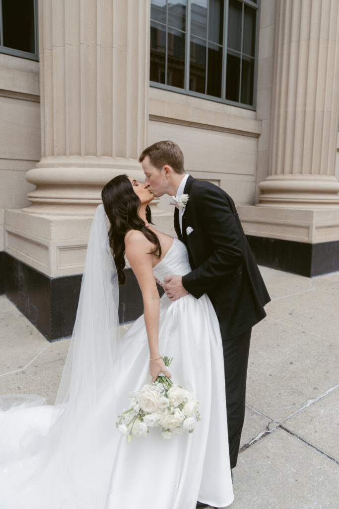 The bride and groom share a kiss outside a grand building on their wedding day.