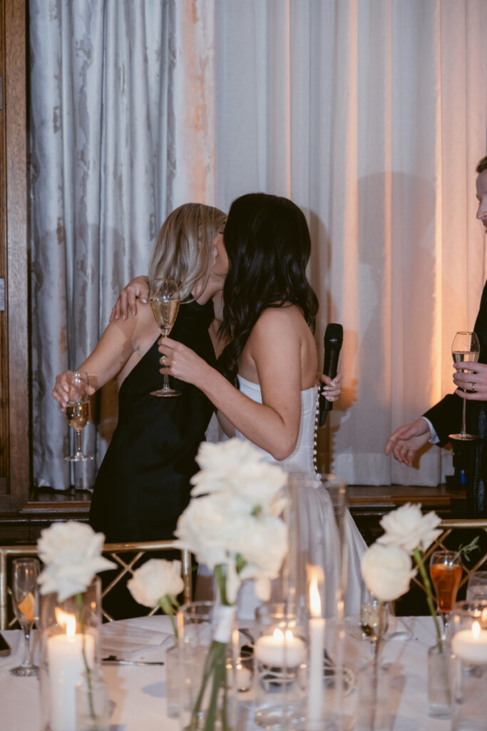 The bride hugs a bridesmaid with champagne in hand after an emotional speech at the reception.