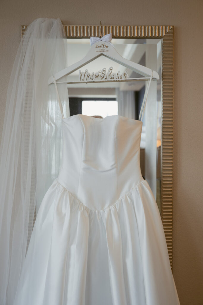 Strapless satin wedding dress hanging on mirror before cozy Christmas wedding in Cincinnati hotel room.