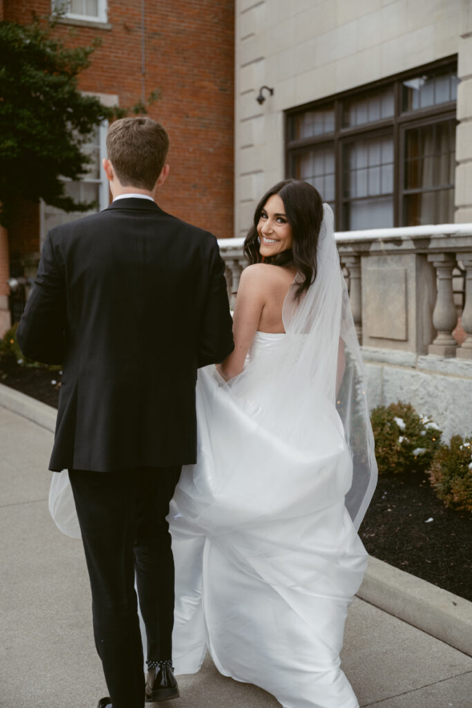 Bride glances back with a smile while walking hand in hand with the groom outside.