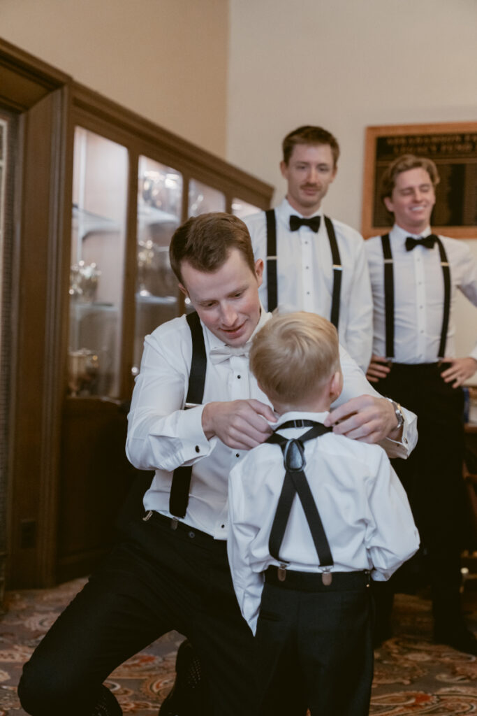 Groom helping ring bearer with bow tie before cozy Christmas wedding, surrounded by groomsmen in suspenders.