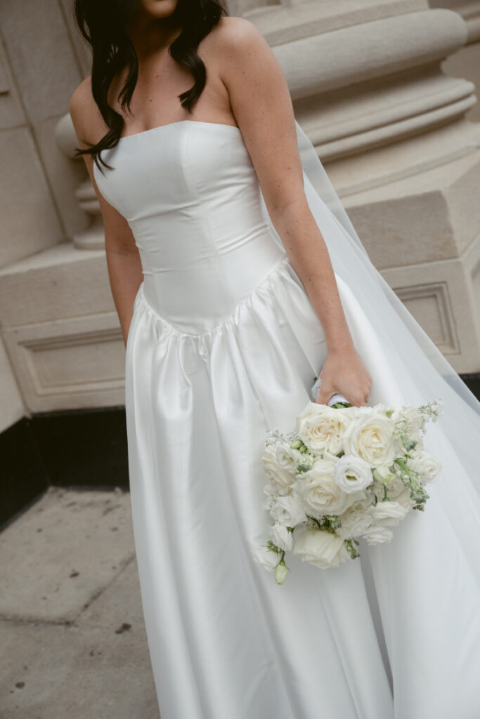 Bride holding a white rose bouquet, standing outside in her strapless satin wedding dress.