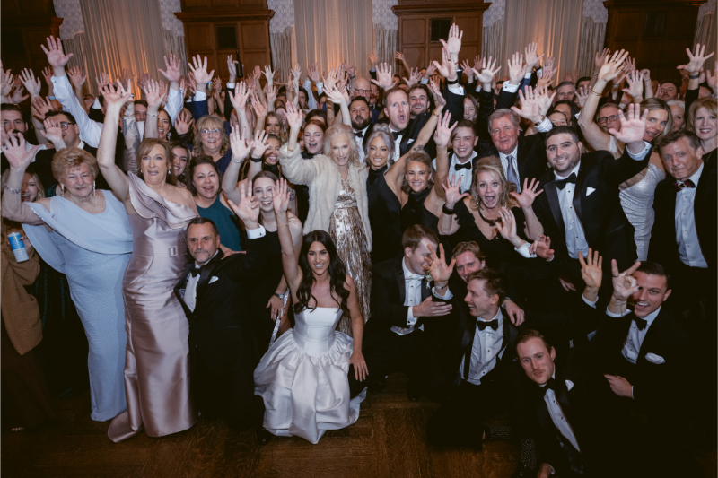 The bride poses with a joyful crowd of wedding guests, all raising their hands in celebration.