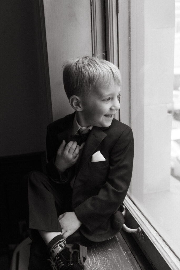 Ring bearer in suit smiling while sitting on windowsill before cozy Christmas wedding ceremony.